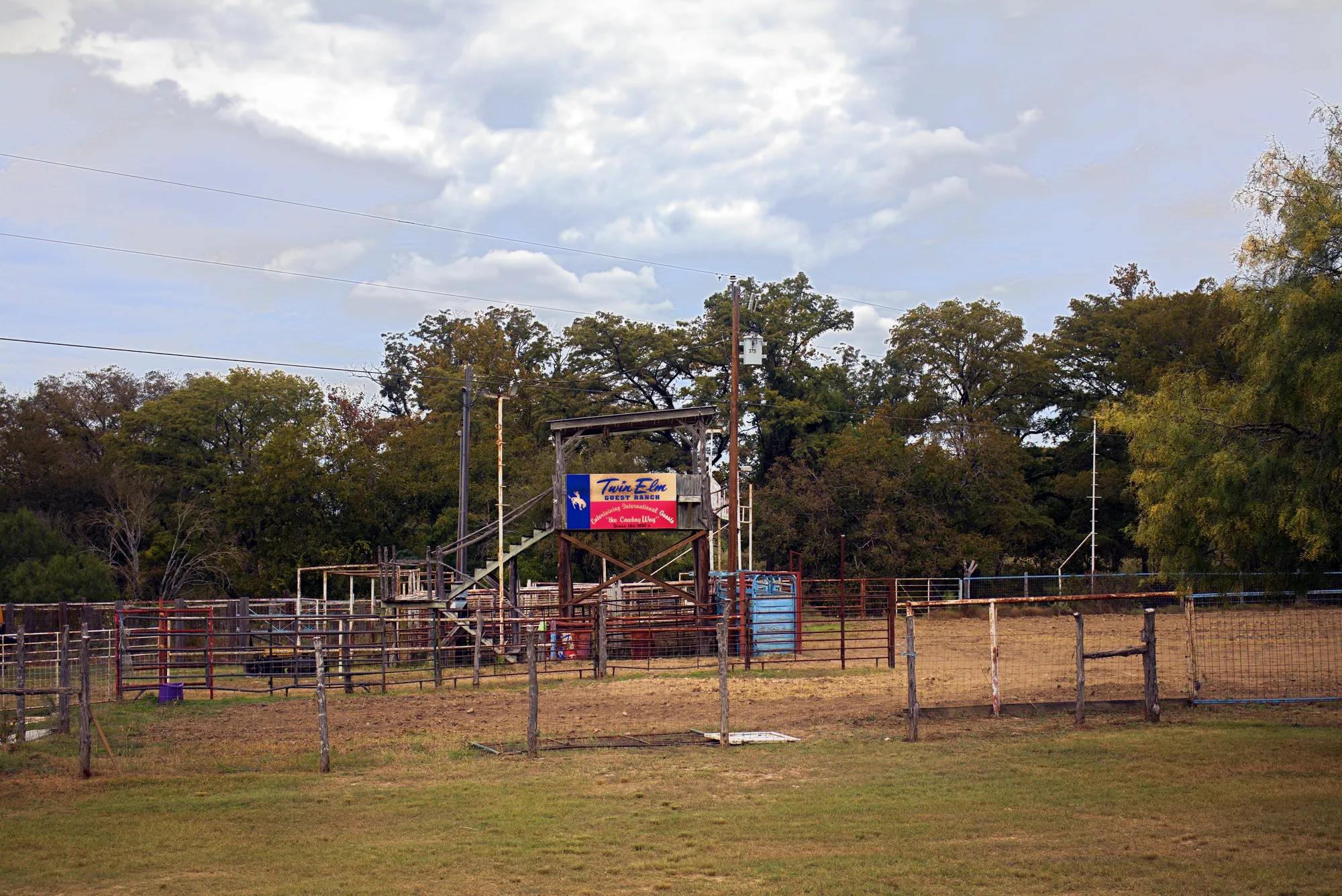 The Twin Elm rodeo arena: pens and chutes, the announcer stand with its Twin Elm Guest Ranch banner, and big oaks behind under a cloudy sky