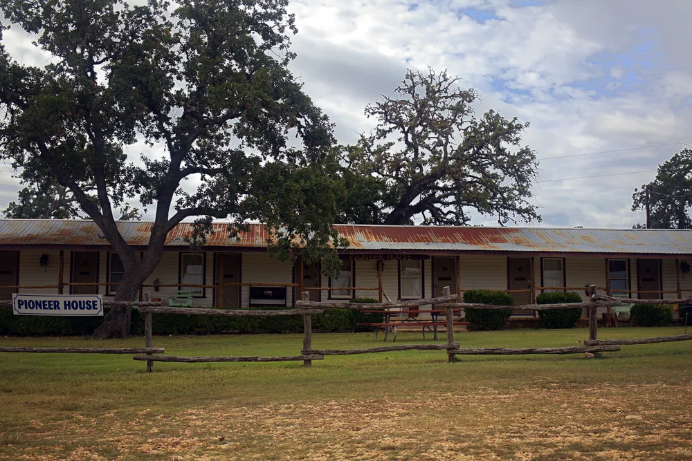 The Pioneer House, a long white building with a tin roof and a covered porch under big live oaks, its sign on the cedar rail fence out front