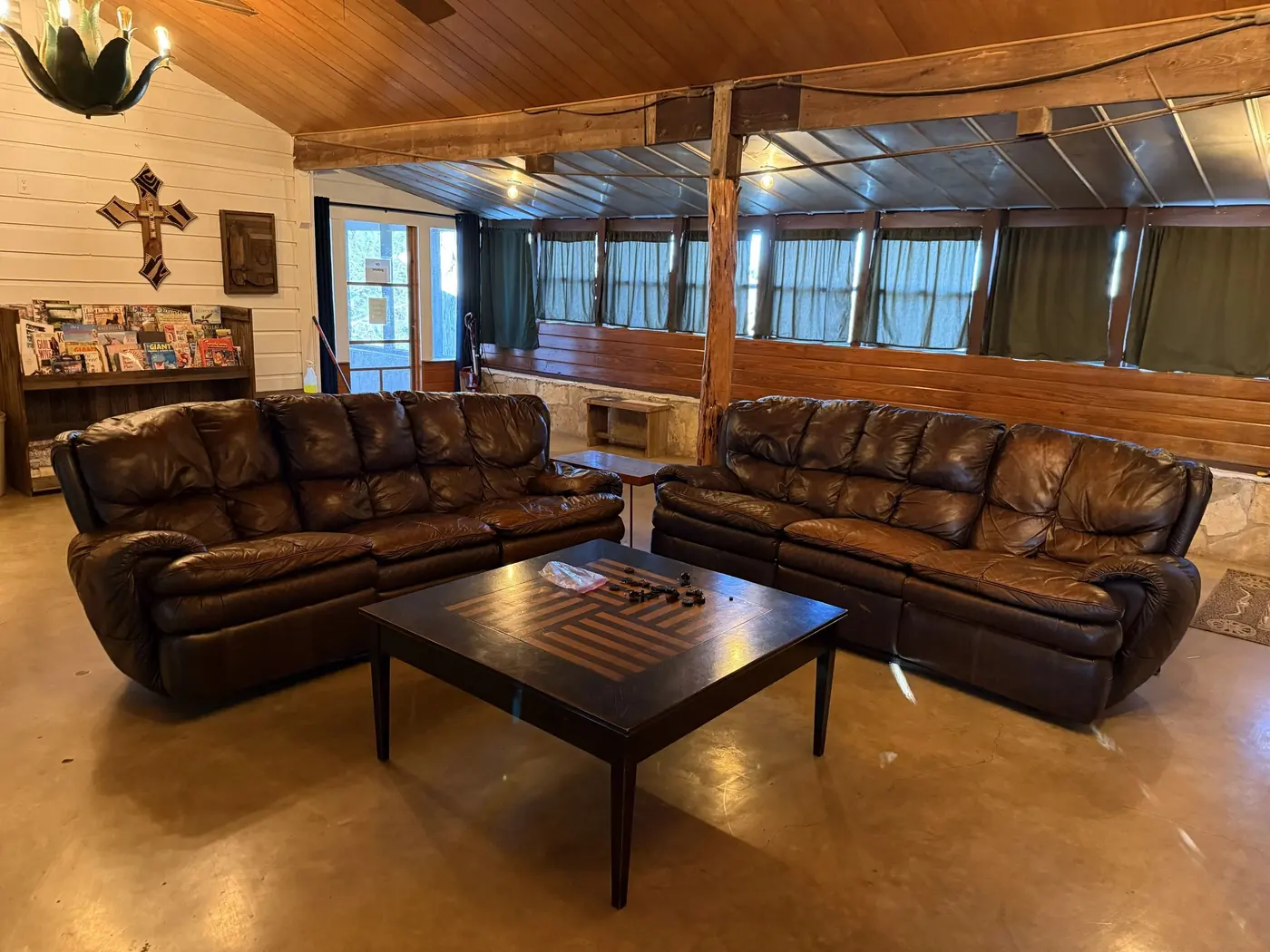 A common room with two leather sofas around a checkerboard coffee table, a brochure rack and a long wall of curtained windows