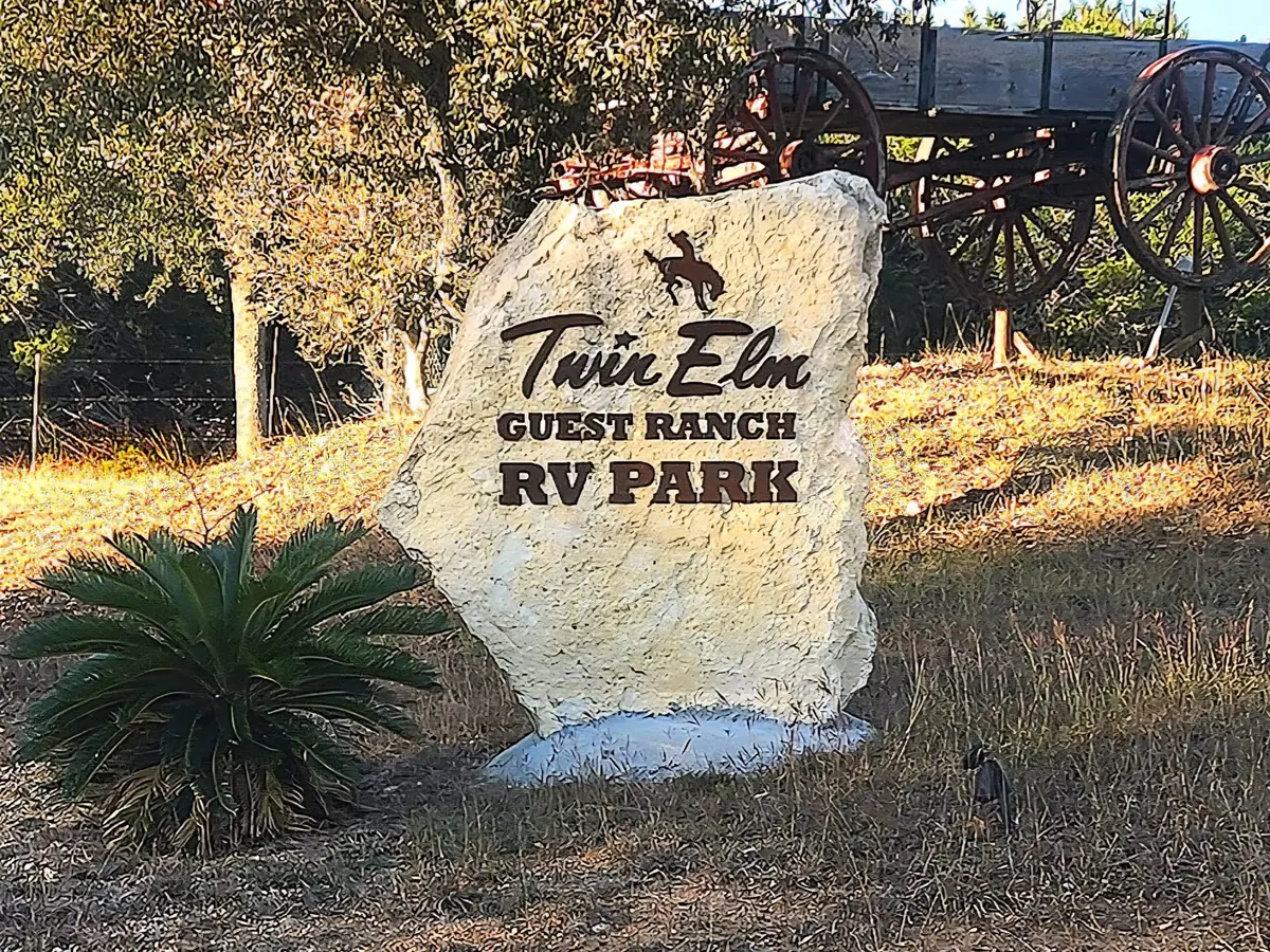 The carved limestone Twin Elm Guest Ranch RV Park sign in late sun, with an old wagon behind it