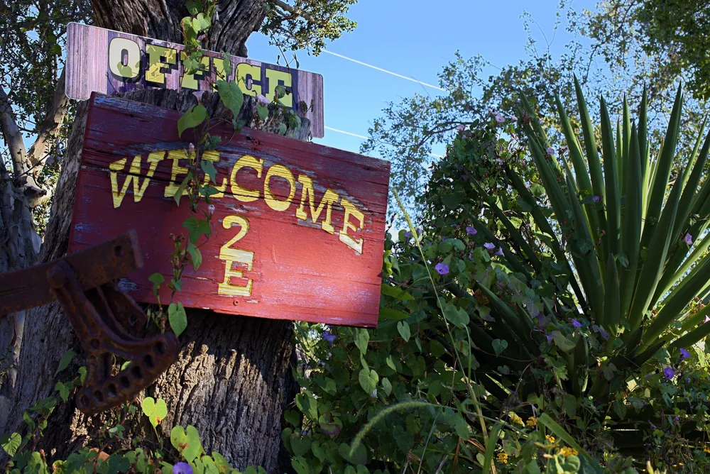 A red &ldquo;Welcome&rdquo; sign with the 2E brand under an &ldquo;Office&rdquo; sign, nailed to a tree beside a yucca